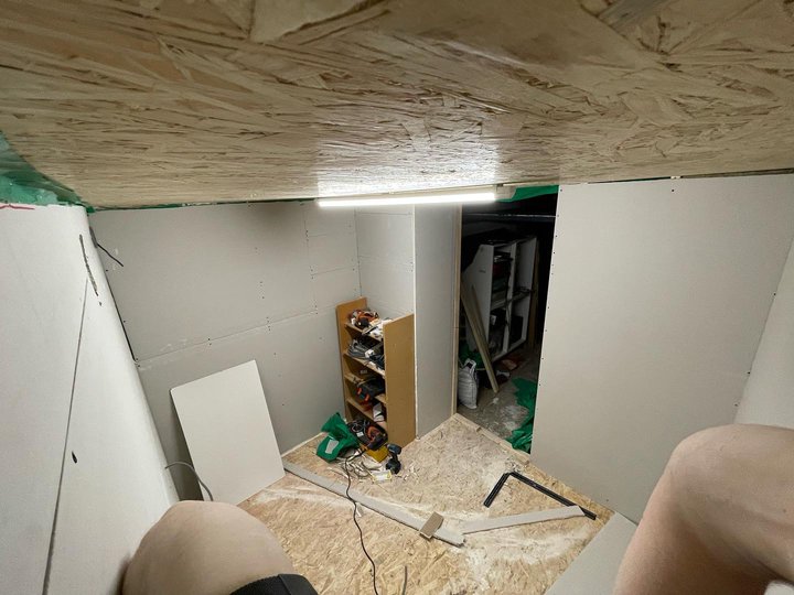 A corner shot looking towards a bare doorway, the walls are covered with bare plasterboard. A shelf holding tools is next to the doorway.
